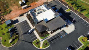 Aerial view of a new commercial building with top roofing colors, featuring a large parking lot filled with several cars and landscaped areas.